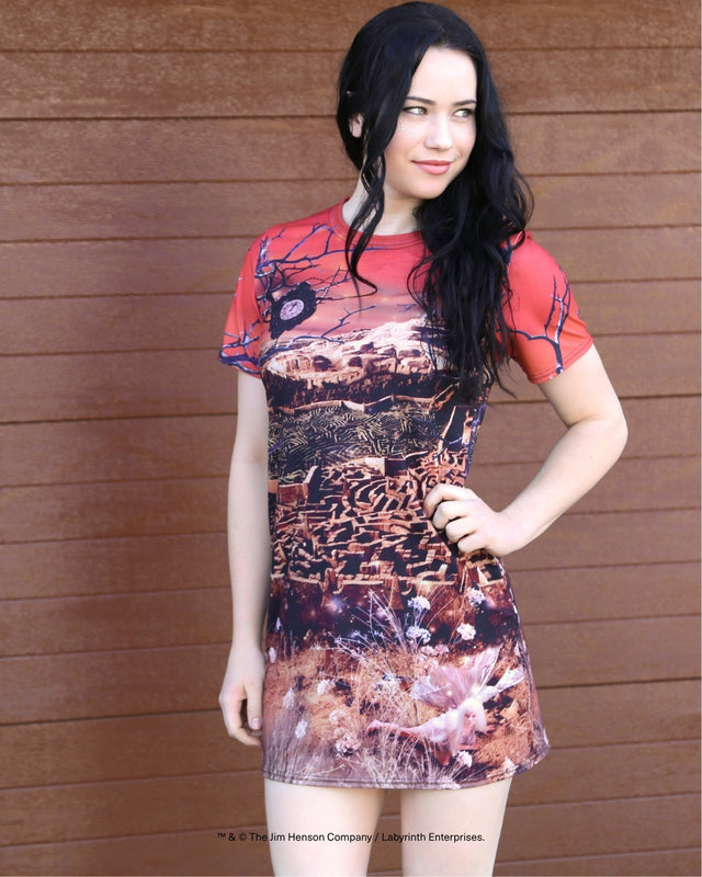 A woman modeling a short-sleeve t-shirt dress with a red crew neck and a detailed western landscape print in brown, beige, and black, posed against a brown wooden wall.