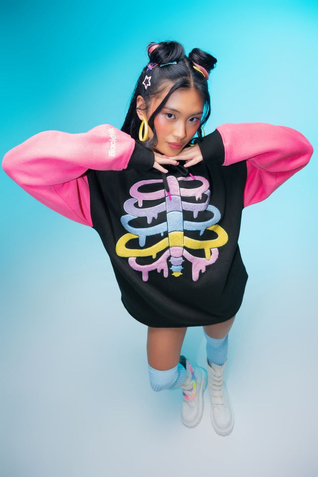 Oversized black fleece hoodie featuring a colourful rainbow skeleton ribcage graphic, bright pink sleeves, embroidered BlackMilk logos and a cosy unisex streetwear fit.