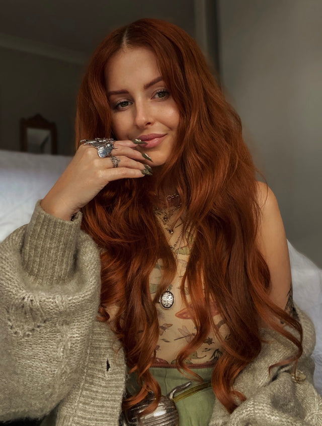 A woman with long, wavy auburn hair is shown in a close-up portrait, wearing a chunky knit beige cardigan, a patterned top, and multiple silver rings. She is resting her chin on her hand in an indoor setting.