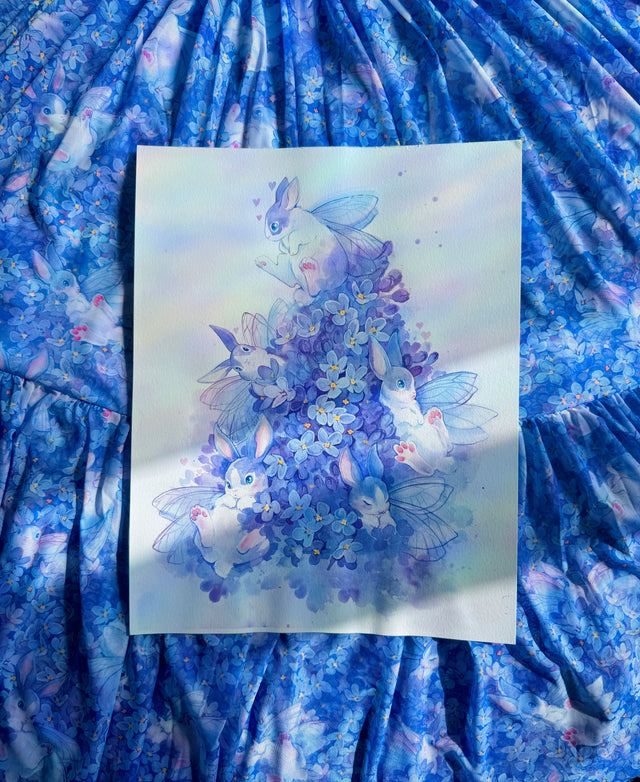 Artistic depiction of fairies and flowers on a white card, placed on a blue floral fabric background.