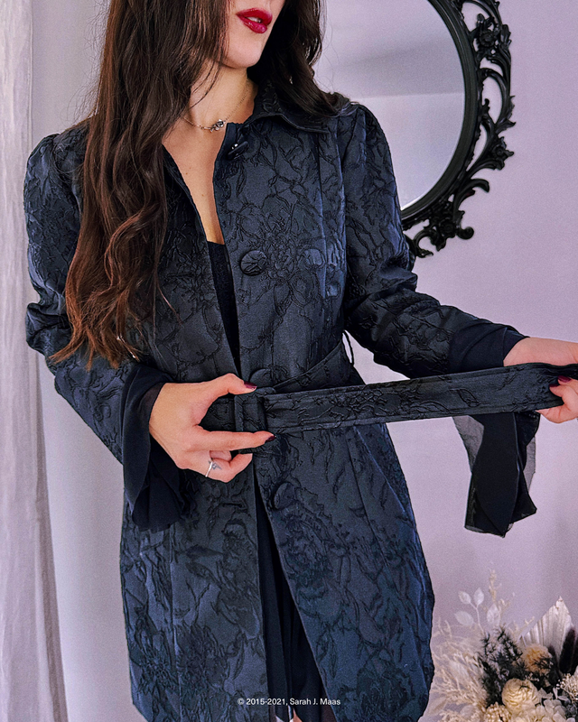 Model wearing a dark, textured jacket with a subtle floral jacquard design, tying the waist belt to create a fitted silhouette, inspired by the romantic, moody world of A Court of Thorns and Roses (ACOTAR).