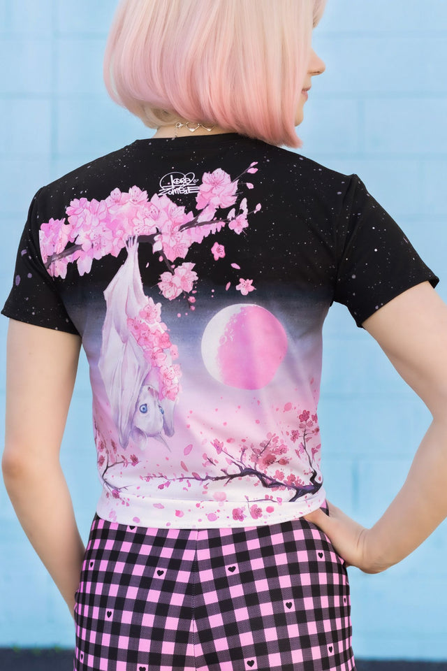 Person wearing a black t-shirt with a colorful design of a bat, cherry blossoms, and a moon against a blue background.