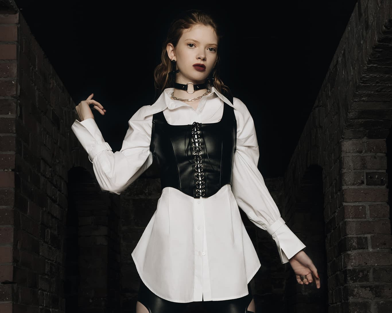 Person wearing a black leather outfit with a white shirt against a dark brick wall.