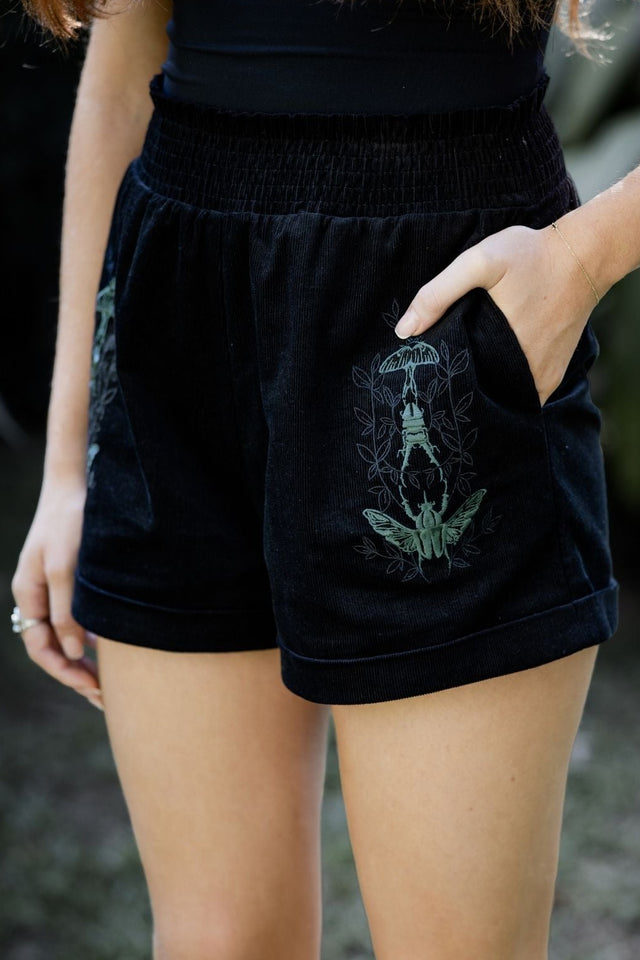 Fantasy witch inspired black corduroy shorts embroidered with deep green moth artwork by Ami Raithe. Featuring a relaxed fit, cuffed hems and elasticated paper bag waist, these goth aesthetic shorts bring together whimsigoth, alternative fashion and dark cottagecore styling from the Nature of a Witch collection.