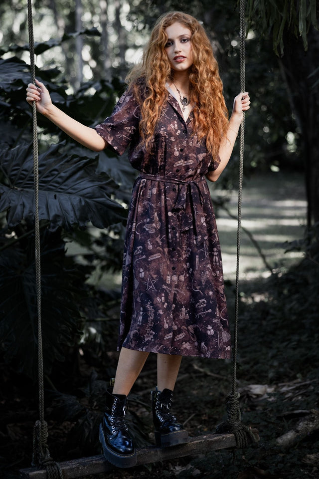Witchy button-through midi dress featuring Ami Raithe artwork with potion bottles, bones, butterflies and mystical apothecary details scattered across a rich earthy print. This whimsigoth dress blends cottage witch, dark academia and fantasy witch fashion with a floaty A-line silhouette and vintage-inspired styling.
