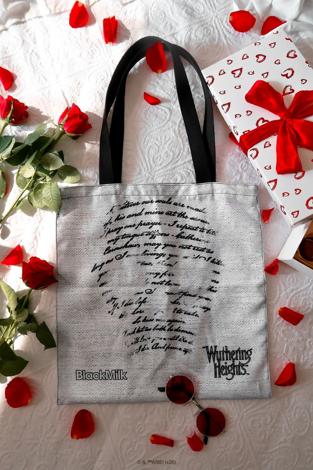 Drive Me Mad Tote Bag from the Wuthering Heights collection, a gothic grey fabric tote featuring handwritten script text and BlackMilk branding, styled flatlay with red rose petals