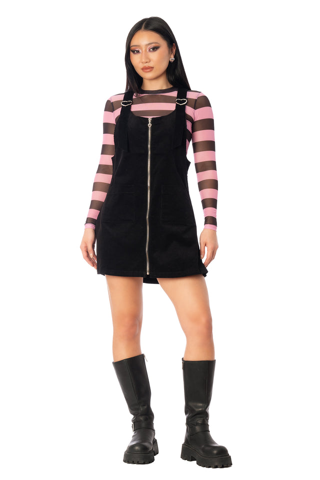 Model wearing the black cord pinny dress over a pink-and-black striped top. League of Legends K/DA x BlackMilk black corduroy pinafore mini dress with shoulder straps and a straight-cut silhouette.