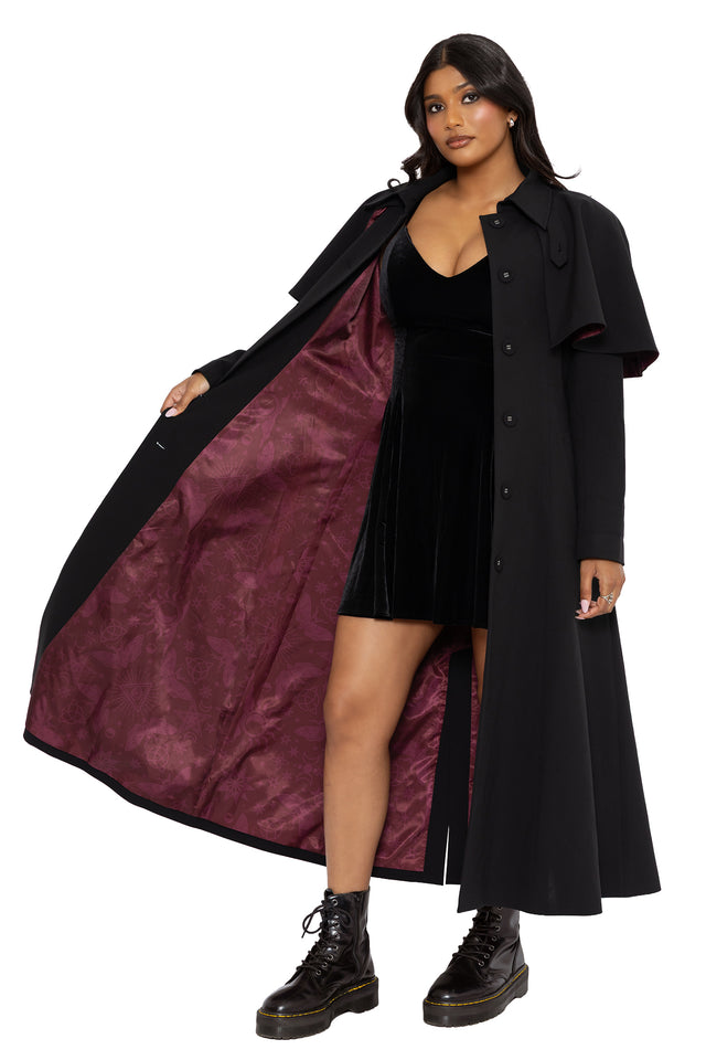 Alternative witchcore trench coat with a structured black silhouette and hidden spell inspired lining for subtle gothic styling. This long gothic coat blends whimsigoth and corporate goth aesthetics with a removable cape detail and elegant full-length design.